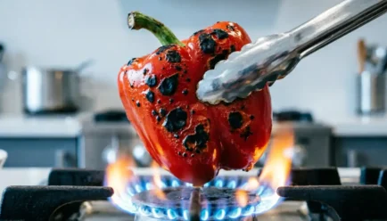 : Metal tongs holding a blistered, charred red bell pepper directly over a blue gas burner flame.