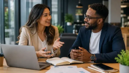 Two diverse professionals having a friendly discussion in a modern office with a laptop, notebook, smartphone, documents, and workspace materials.