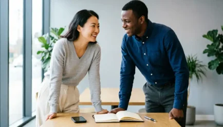 Two diverse professionals standing at a clean wooden desk in a modern office, smiling and engaging in a warm conversation. A smartphone, open notebook, and pen are on the desk.