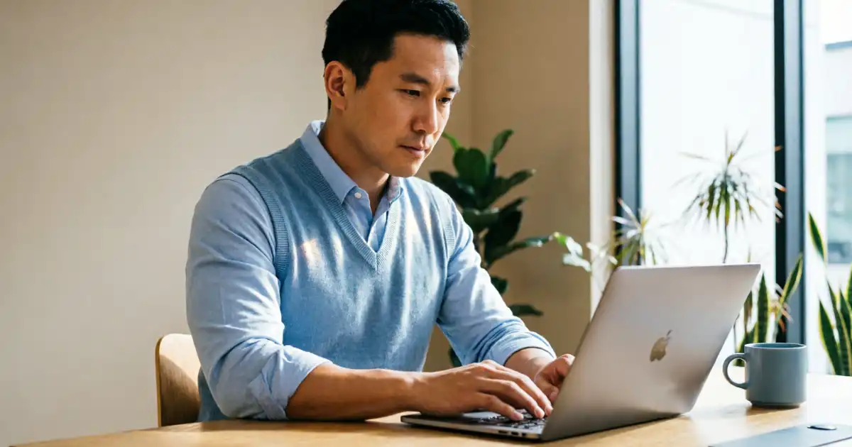 Professional person typing on laptop computer at wooden desk in bright minimalist office with large windows, soft natural lighting, neutral beige walls, indoor plants, and soft blue accents, clean modern workspace