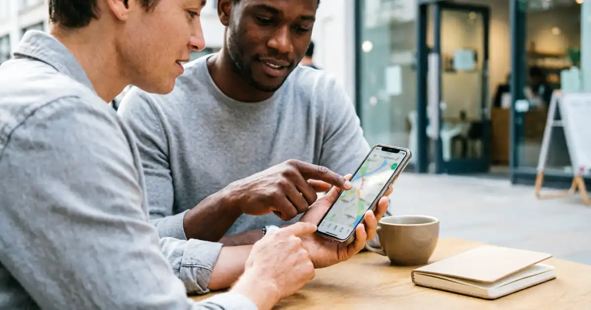 Two diverse adults sitting at an outdoor cafe table, looking at a map on a smartphone to find nearby locations, with a coffee cup and notebook on the table and blurred urban storefronts in the background.