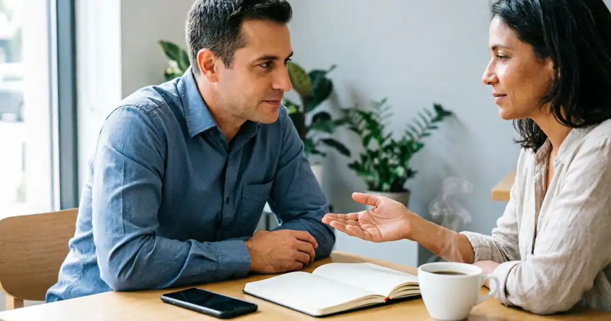 Two diverse adults sitting at a wooden table in a bright cafe, one person listening attentively while the other speaks with an open hand gesture, with a smartphone, open notebook, and coffee cup on the desk.